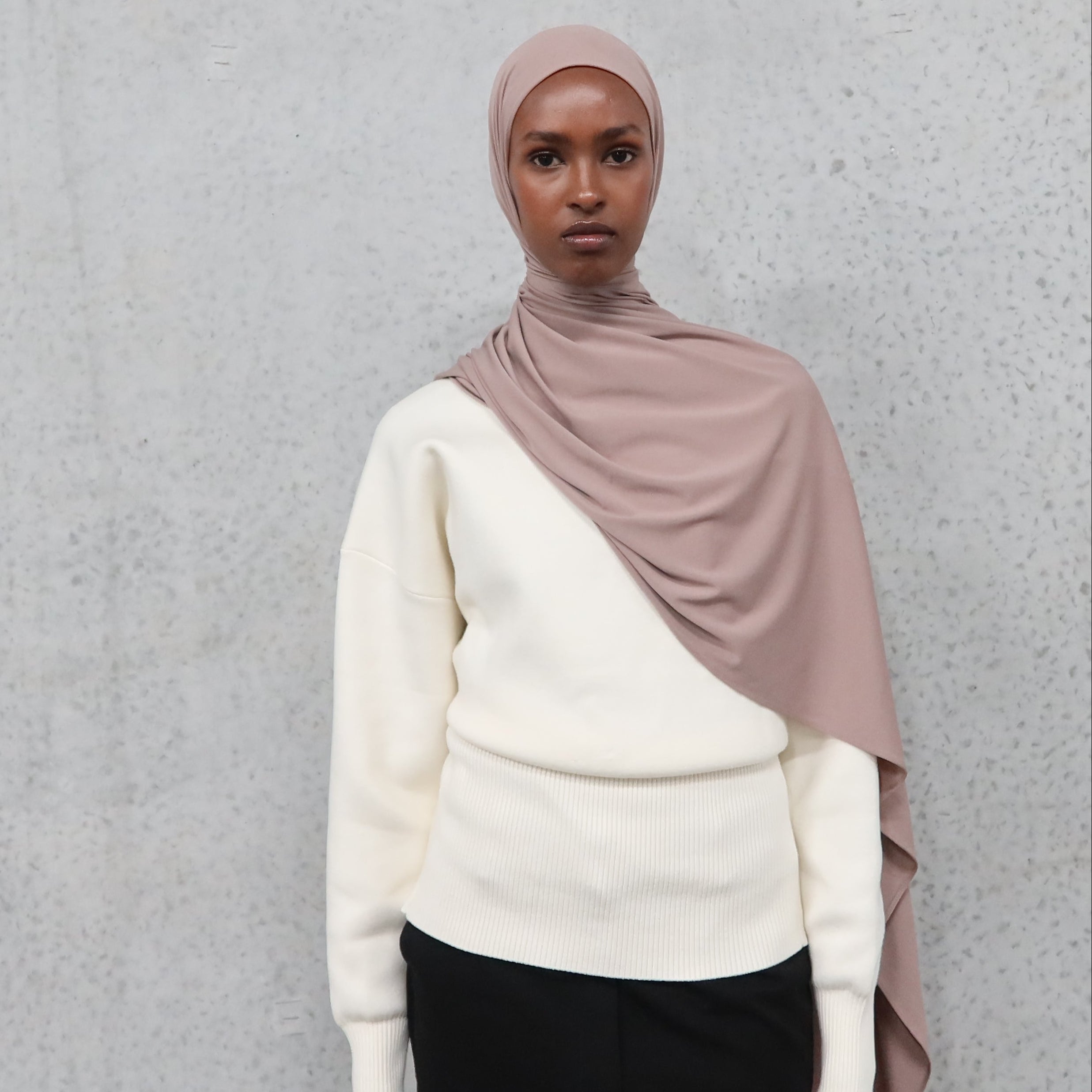 Person wearing a beige hijab and white sweater against a gray background